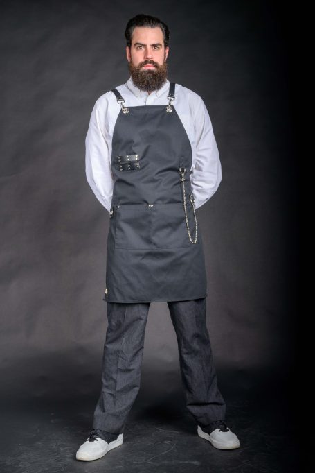 Elegant black polycotton apron with cross-back leather straps, central pocket, and metal accents. Versatile and comfortable for professionals like baristas, chefs, and bartenders. Thoughtful design with divided waist pocket for functionality.