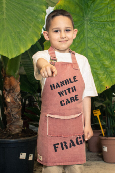 Trendy stonewashed denim kids' apron in light maroon color, with adjustable neck strap and functional pocket. Street style vibes and hand-painted details.