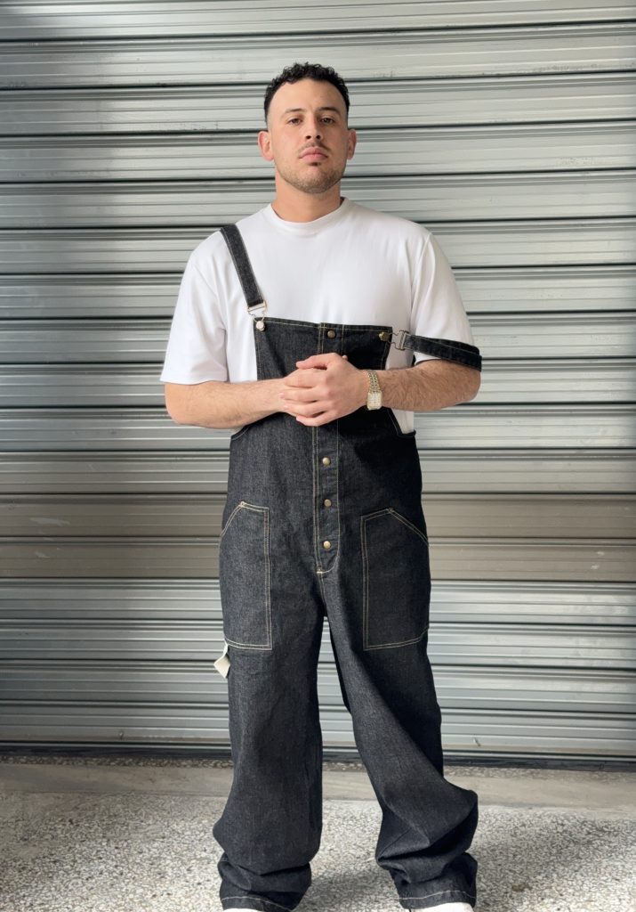 Man wearing a stylish black denim overall with a white T-shirt, standing in front of a metallic background, showcasing the front view of the overall.