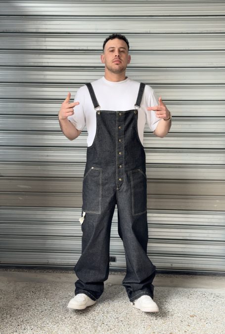 Man wearing a dark denim overall paired with a white T-shirt, standing confidently in front of a metallic shutter background, showcasing the front design and fit