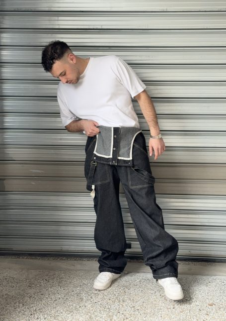 Man posing with his hands in the pockets of a black denim jumpsuit, paired with a white T-shirt, in front of an industrial backdrop.