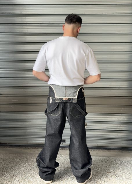 Back view of a black denim dungaree worn by a man with a white T-shirt, standing against a metallic surface.