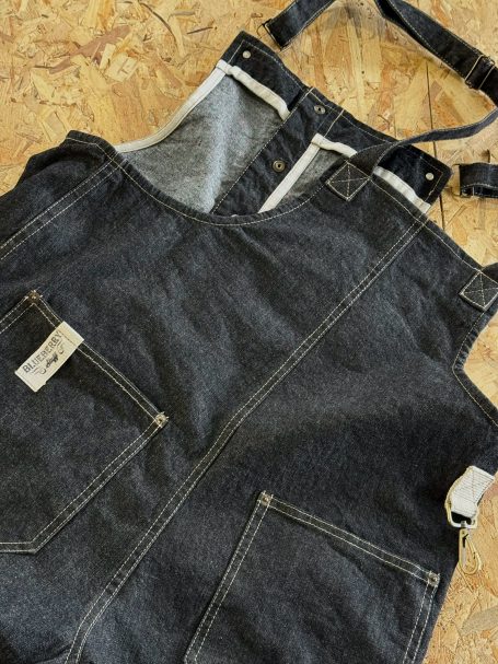 Close-up of a black denim bib-and-brace garment laid on a wooden surface, featuring visible stitching and metallic hardware details.