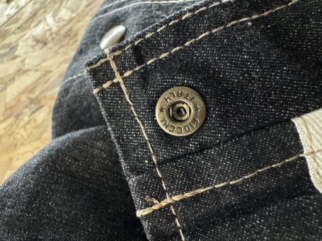 Close-up of a black denim bib-and-brace garment laid on a wooden surface, featuring visible stitching and metallic hardware details.